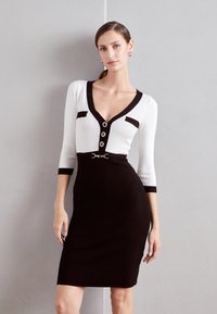 Robe en tricot avec une palette de couleurs noir et blanc, décolleté en V profond, manches trois-quarts, accents noirs et quincaillerie argentée à la taille.