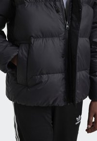 Black puffer jacket with glossy finish, zip closure, and snap buttons. Features two front pockets and a slightly padded texture.