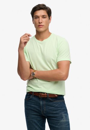 Jonge man die een lichtgroen shirt met korte mouwen, donkere jeans, een bruine riem en een polshorloge draagt, poseert met één arm gekruist en de andere omhoog.