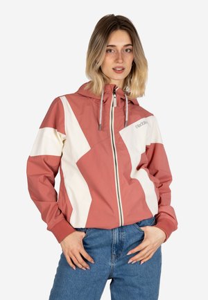 Young woman with shoulder-length hair wearing a pink and white zip-up jacket with hood and blue jeans, standing with hands in pockets.