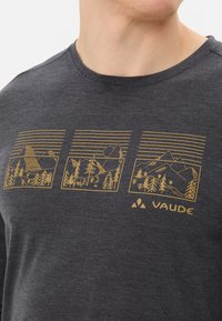Donkergrijze T-shirt met ronde hals featuring gouden berglandschapontwerpen in drie rechthoekige kaders. Merklogo "VAUDE" aan de onderkant.