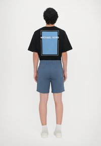 Tricou negru cu mânecă scurtă, având un grafic rectangular albastru și textul "MICHAEL KORS" pe spate, combinat cu pantaloni scurți albaștri și încălțăminte albă.