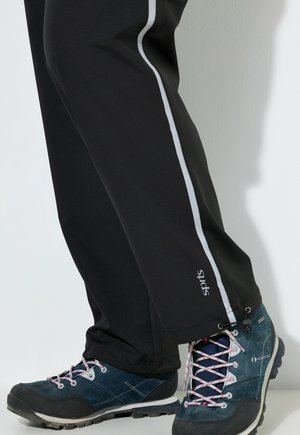 Ben iført sorte sportbukser med hvide sidestriber og blå vandresko med pink og hvide snørebånd stående på en hvid overflade.