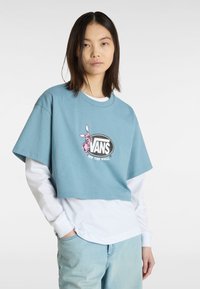 Lyseblå cropped bomulds-t-shirt med et pink kaninmotiv og sort "VANS" logo, lagd ovenpå en hvid langærmet skjorte.