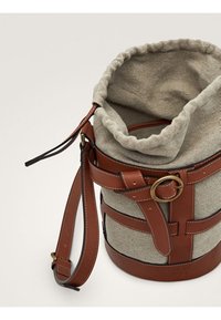 Borsa a secchiello in tessuto beige con manici in pelle marrone, fibbia dorata e tracolla regolabile su uno sfondo chiaro.