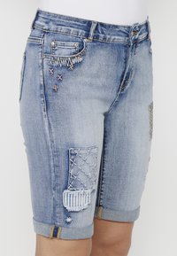 Shorts en denim bleu clair de type bermuda avec divers détails en patchwork, des coutures décoratives et des poignets retroussés agrémentés de quincaillerie.