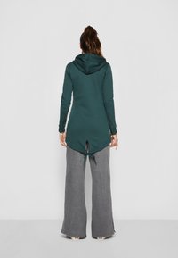 Tunic verde acqua con cappuccio e maniche lunghe, con orlo spaccato e dettaglio con cravatta sul retro, abbinata a pantaloni grigi a gamba ampia.