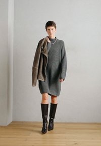 Abito in maglia grigia con maniche lunghe, abbinato a una giacca in suede marrone poggiata su un braccio e stivali alti fino al ginocchio in pelle marrone.