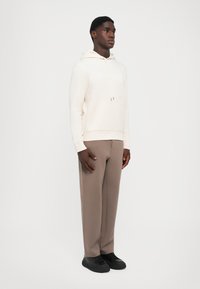 Kremfarget hettegenser med lommer foran, beige bukser og svarte joggesko. Minimalistisk design, myk tekstur og nøytral fargepalett.
