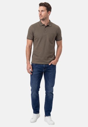 Mann trägt ein olivgrünes Poloshirt, blaue Jeans und weiße Sneakers, steht mit einer Hand in der Tasche und schaut zur Seite.