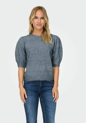 Donna con capelli biondi che indossa un top in maglia grigio con maniche a sbuffo e jeans blu, in piedi di fronte a uno sfondo grigio chiaro.