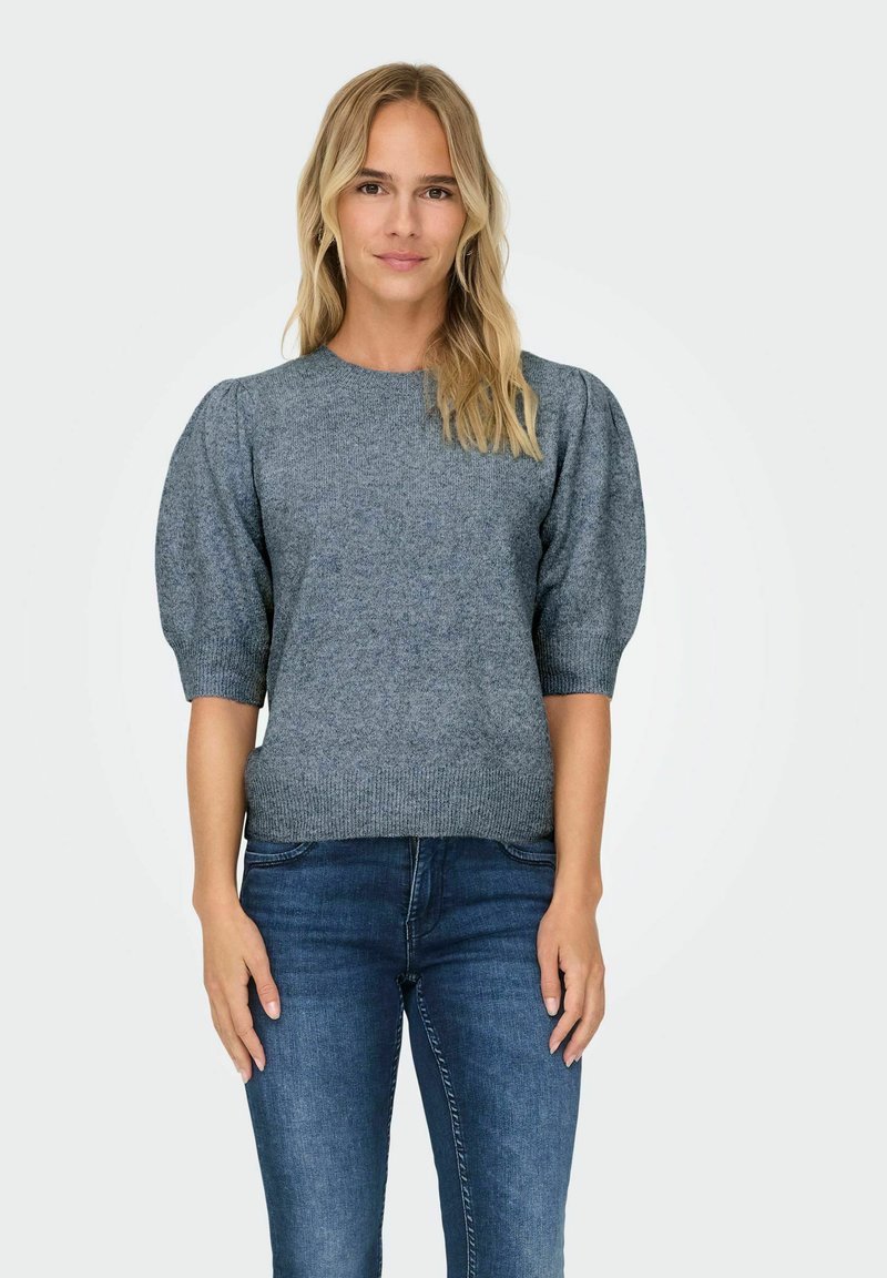 Femme aux cheveux blonds portant un haut en tricot à manches bouffantes grises et un jean bleu, se tenant devant un fond gris clair.