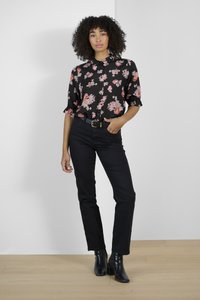 Chemise noire à motifs floraux rouges et blancs, à manches courtes ; associée à un jean noir et des bottines. Le modèle se tient sur un sol en bois.