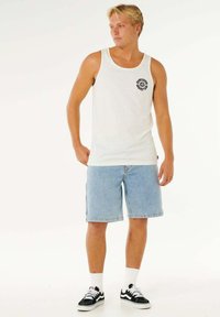 Camiseta sin mangas blanca con un logo negro, combinada con pantalones cortos de mezclilla azul claro. Zapatillas negras con detalles blancos y calcetines blancos.