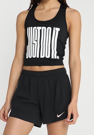 Kobieta nosząca czarny bezrękawnik typu crop top z napisem "JUST DO IT." oraz czarne sportowe spodenki z różowym logo Nike na lewej nodze.