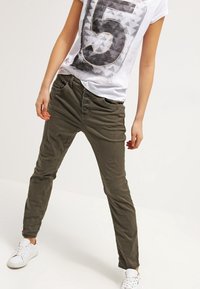 Persona que lleva pantalones de botonera verde oliva, zapatillas blancas y una camiseta blanca con un gran número 5 impreso, de pie con las piernas separadas.