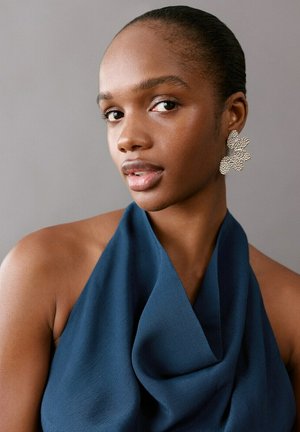 Jeune femme aux cheveux tirés vers l'arrière portant de grandes boucles d'oreilles argentées à motifs floraux et un haut dos nu bleu sur un fond gris uni.