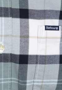 Close-up van een grijs-wit-zwart geruit overhemdzakje met een Barbour merklabel en een witte knoop bij de naad.