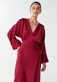 Burgundy satin dress featuring a deep V-neck, long billowy sleeves, and a cinched waist, creating an elegant silhouette with smooth texture.
