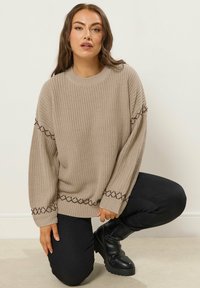 Beige neulepaita, jossa on rento leikkaus. Sen teksturoitu kangas ja kontrastivärinen ruskea tikkaus hihojen ja helman kohdalla tuovat tyyliä. Mustat housut ja saappaat.