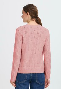 Maglione rosa lavorato a maglia con un motivo testurizzato, maniche lunghe e scollo rotondo; polsini e orlo a coste. Indossato con jeans blu.