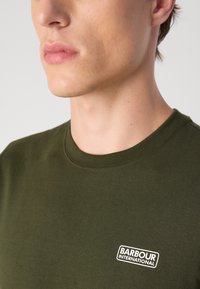 Eλιά πράσινη βαμβακερή μπλούζα T-shirt με στρογγυλή λαιμόκοψη, που διαθέτει λευκή θηλιά λογότυπου "BARBOUR INTERNATIONAL" στην αριστερή περιοχή του στήθους.