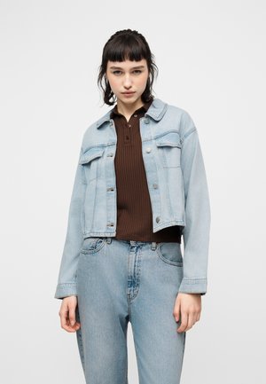 Jeune femme portant une veste en denim bleu clair et un jean avec un polo côtelé marron, debout devant un fond clair uni.