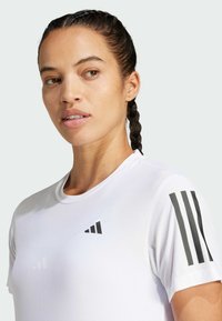 Camiseta deportiva blanca hecha de tejido texturizado, con un logo negro y tres franjas verticales en el hombro izquierdo. Mangas cortas.