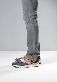 Buty sportowe z mieszanką niebieskiego zamszu i beżowych akcentów, posiadające białą, nakropioną podeszwę i szczegółowe sznurowanie. Noszone z szarymi, dopasowanymi dżinsami.