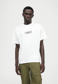 Biela bavlnená tričko s okrúhlym čiernym logom na hrudi, s uvoľneným strihom a krátkymi rukávmi. Nosí sa s olivovozelenými cargo nohavicami.