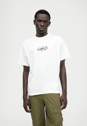 Hvit bomull t-skjorte med et sirkulært svart logo på brystet, med en avslappet passform og korte ermer. Brukt med olivengrønn cargo-bukse.