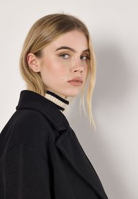 Schwarzer Mantel mit Reverskragen, aus strukturiertem Stoff. Darunter ein schwarz-weiß gestreifter Rollkragenpullover. Das Model hat schulterlanges blondes Haar.