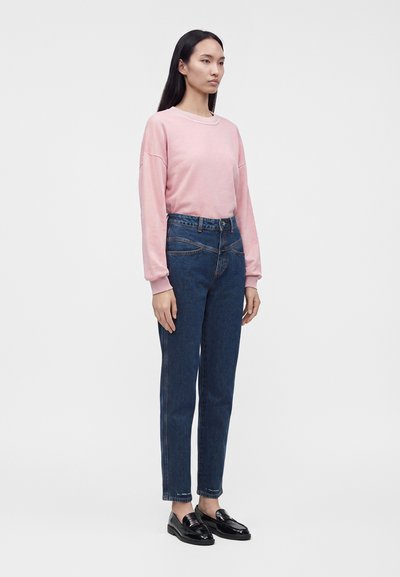 Femme debout portant une chemise rose à manches longues, un jean bleu taille haute et des mocassins noirs sur un fond clair uni.