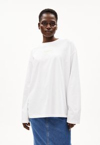 Camicia bianca a maniche lunghe con colletto rotondo, con la scritta "ARMEDANGELS" in caratteri dorati. Indossata con una gonna in denim blu, dal tessuto liscio.