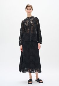 Černý krajkový outfit skládající se z dlouhého rukávu, button-up topu a odpovídající maxi sukně, obojí s složitými vzory, doplněný černými sandály.