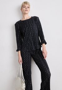 Ensemble noir à motif composé d'un haut à manches longues et d'un pantalon ample, avec un design subtil, un tissu texturé et des poignets à volants. Tient un sac léger.