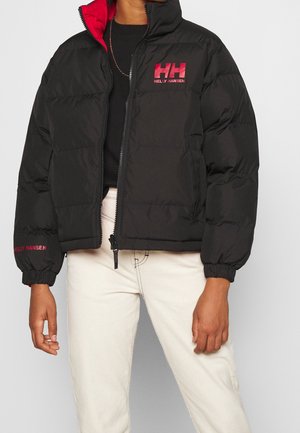Persona con una chaqueta acolchada negra Helly Hansen con logo rojo y pantalones blancos, de pie frente a un fondo liso.