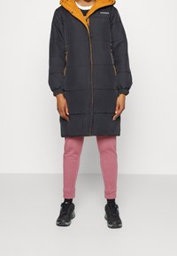 Abrigo negro acolchado con capucha y ribetes naranjas, con cremallera frontal y bolsillos laterales. Se lleva con pantalones deportivos rosas y zapatillas negras.