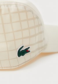 Ljusbeige keps med rutmönster, tillverkad av texturerat material. Har en grön Lacoste-krokodil logotyp med röda accenter.