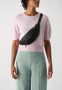 Marsupio in pelle nera con due scomparti con cerniera e tracolla regolabile, indossato sopra un maglione lavorato a maglia rosa chiaro e pantaloni a righe verdi.