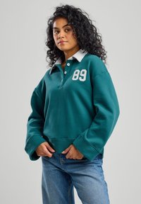 Pullover turchese con collo grigio, caratterizzato da un grande '89' bianco sul petto. Tessuto morbido, vestibilità comoda e maniche lunghe. Design casual.