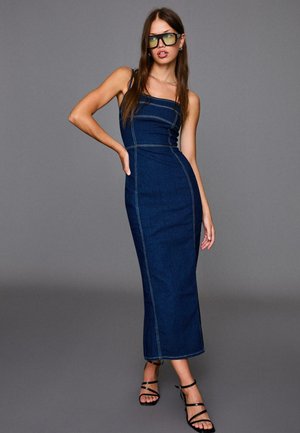 Denim maxi-jurk met een aansluitende lijfje, voorzien van contrasterende stiksels, schouderbanden en een rechte zoom. Gekozen met zwarte sleehakken.