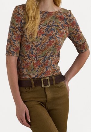 Femme portant un haut à manches courtes à motif floral, rentré dans un pantalon taille haute couleur olive avec une ceinture marron foncé et une boucle carrée décorative.
