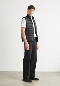 Schwarze Steppweste mit Reißverschluss, leicht und mit genoppter Textur, über einem grauen T-Shirt getragen. Kombiniert mit schwarzen Hosen und Schuhen.