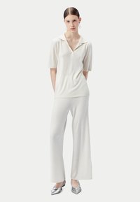 Kvinna står i vit kortärmad topp med krage, ribbade vida byxor, silverfärgade spetsiga skor och stora ringörhängen mot vit bakgrund.