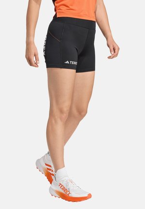 Person, die schwarze Terrex Sportshorts, ein orangefarbenes Oberteil, weiße Socken und weiße sowie orange Terrex Laufschuhe trägt, steht vor weißem Hintergrund.