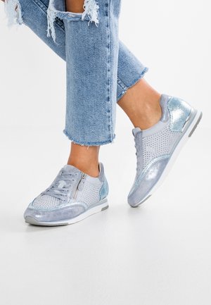Jasnoniebieskie metaliczne trampki z bocznym zamkiem błyskawicznym i perforowanymi detalami, noszone z podartymi, krótkimi niebieskimi jeansami na białej podłodze.