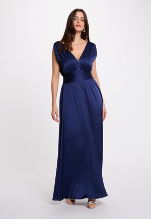 Nainen pukeutuneena hihattomaan, syvän siniseen satiiniseen maksimekkoon, jossa on leveä vyötärönauha ja V-pääntie, yhdistettynä hopeisiin avokkaisiin, seisomassa valkoista taustaa vasten.