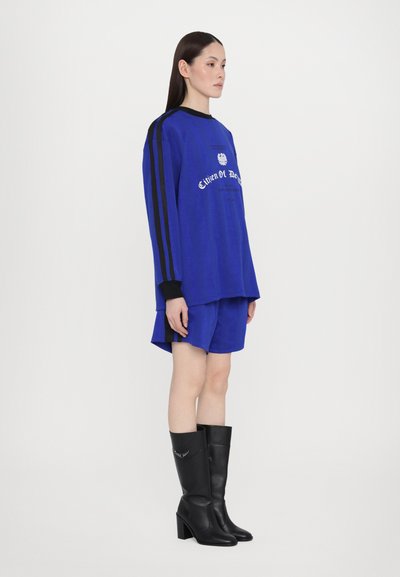 Modra oversized trenerka z črnimi črtami, ki ima natisnjen tekst; v kombinaciji z ustreznimi modrimi kratkimi hlačami in sijočimi črnimi škornji s peto do kolen.