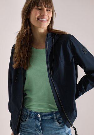 Uśmiechnięta kobieta o długich brązowych włosach, ubrana w granatową kurtkę na zamek, jasnobeżowy t-shirt i niebieskie dżinsy na neutralnym tle.